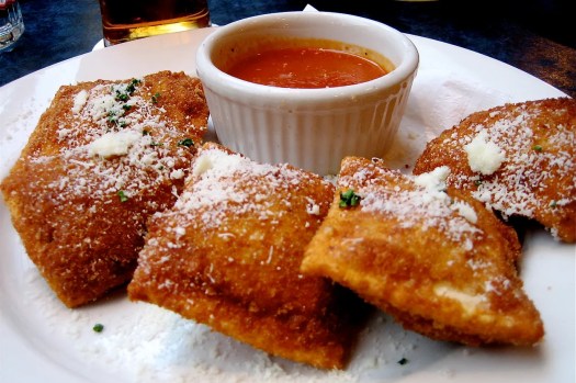 A plate of golden-brown fried ravioli topped with grated cheese and garnished with herbs, served with a side of marinara sauce in a small bowl.